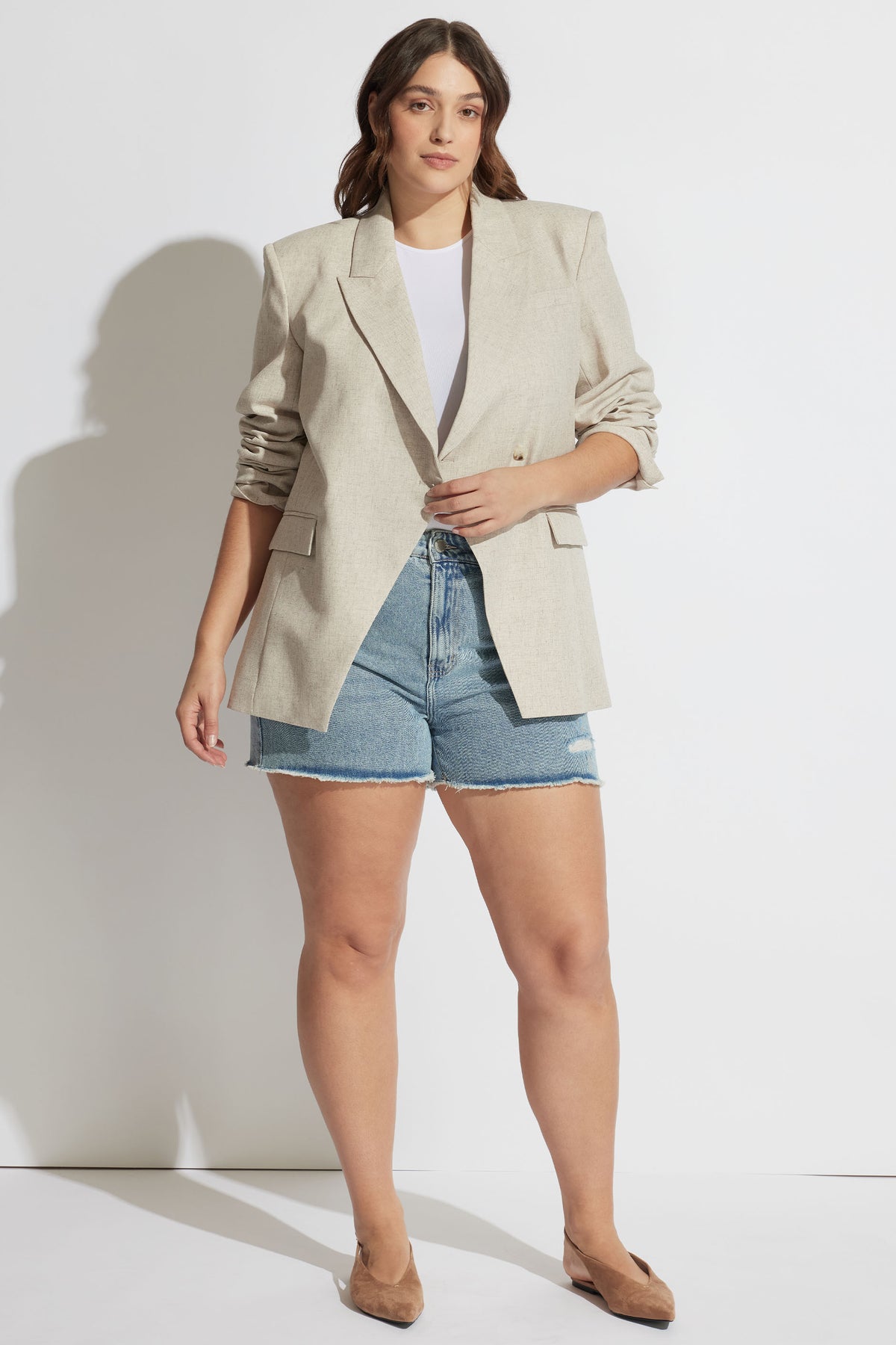 Woman wearing light beige tailored blazer with rolled sleeves paired with blue distressed denim shorts and tan heels