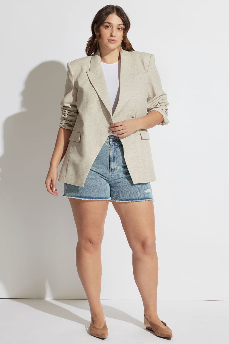Woman wearing light beige tailored blazer with rolled sleeves paired with blue distressed denim shorts and tan heels