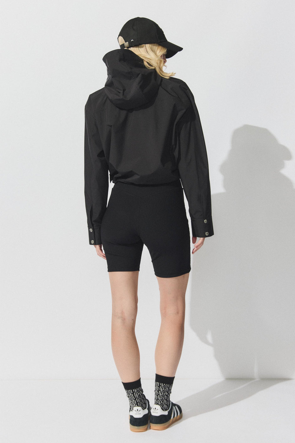 Back view portrait of model wearing black D-Luxe Cropped Spray Jacket with hood, gallery image 3, size 1880x2820 pixels.