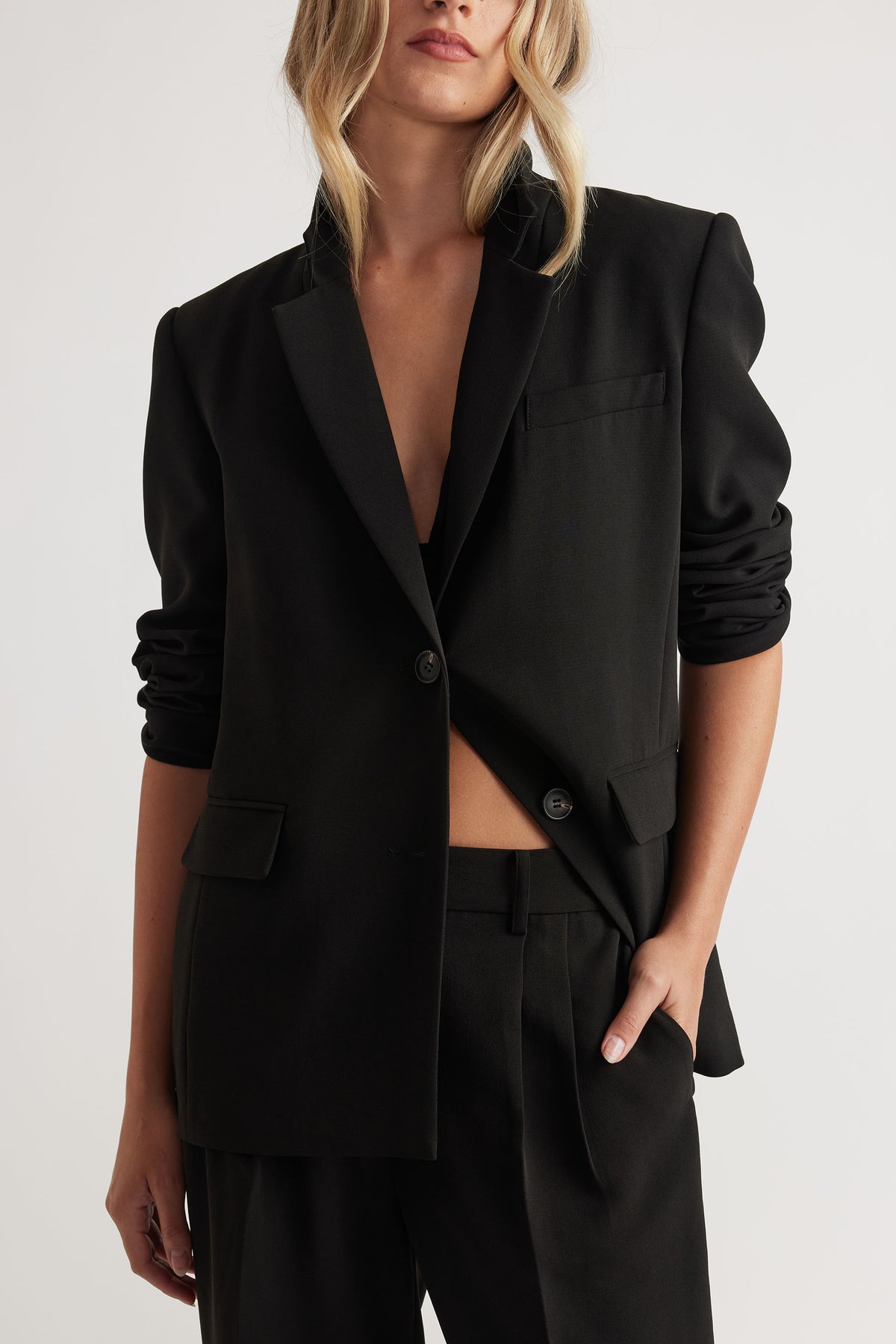 Woman wearing a black tailored blazer with ruched sleeves paired with matching high-waisted trousers and hand in pocket