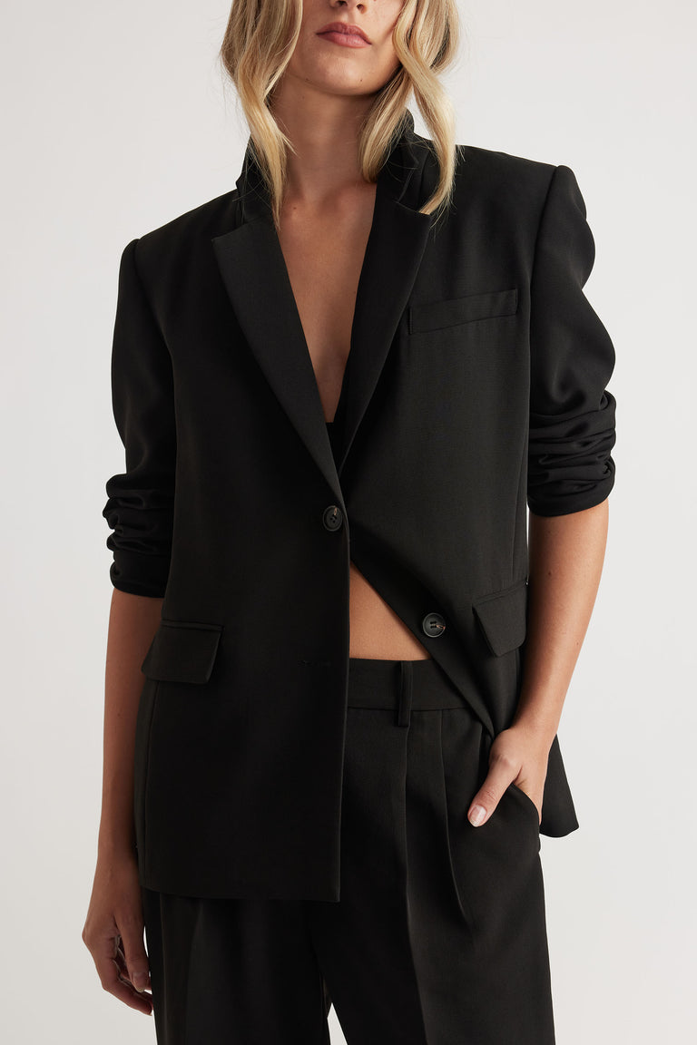 Woman wearing a black tailored blazer with ruched sleeves paired with matching high-waisted trousers and hand in pocket