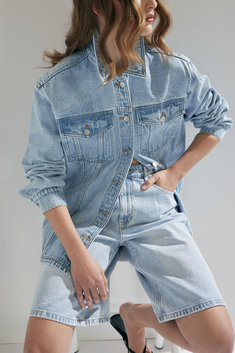 Woman wearing light blue oversized denim jacket and matching denim shorts, with wavy hair and relaxed pose