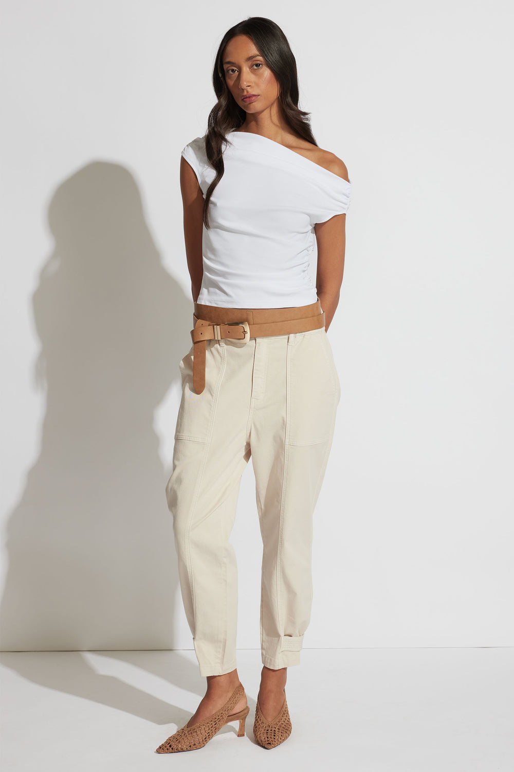 Woman wearing an off-shoulder white top paired with light beige pull-on pants and a tan belt, styled with woven heels.
