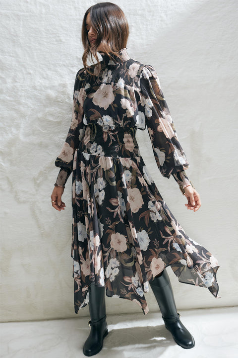 Woman wearing a black floral midi dress with shirred bodice and long sleeves paired with black boots against a white wall.