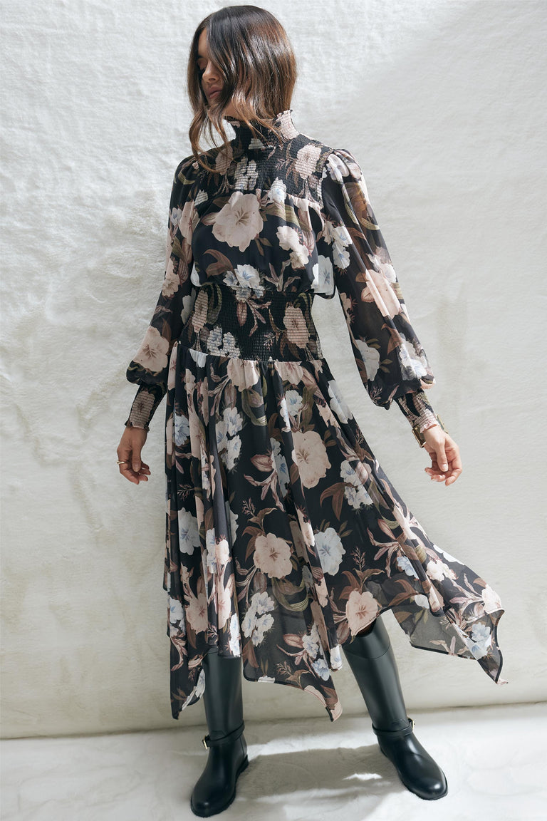 Woman wearing a black floral midi dress with shirred bodice and long sleeves paired with black boots against a white wall.