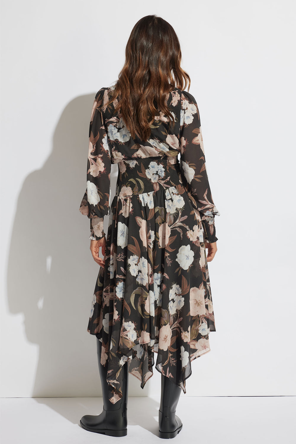 Woman with loose waves wearing a floral midi dress with shirred waist and sheer long sleeves paired with black boots, back view