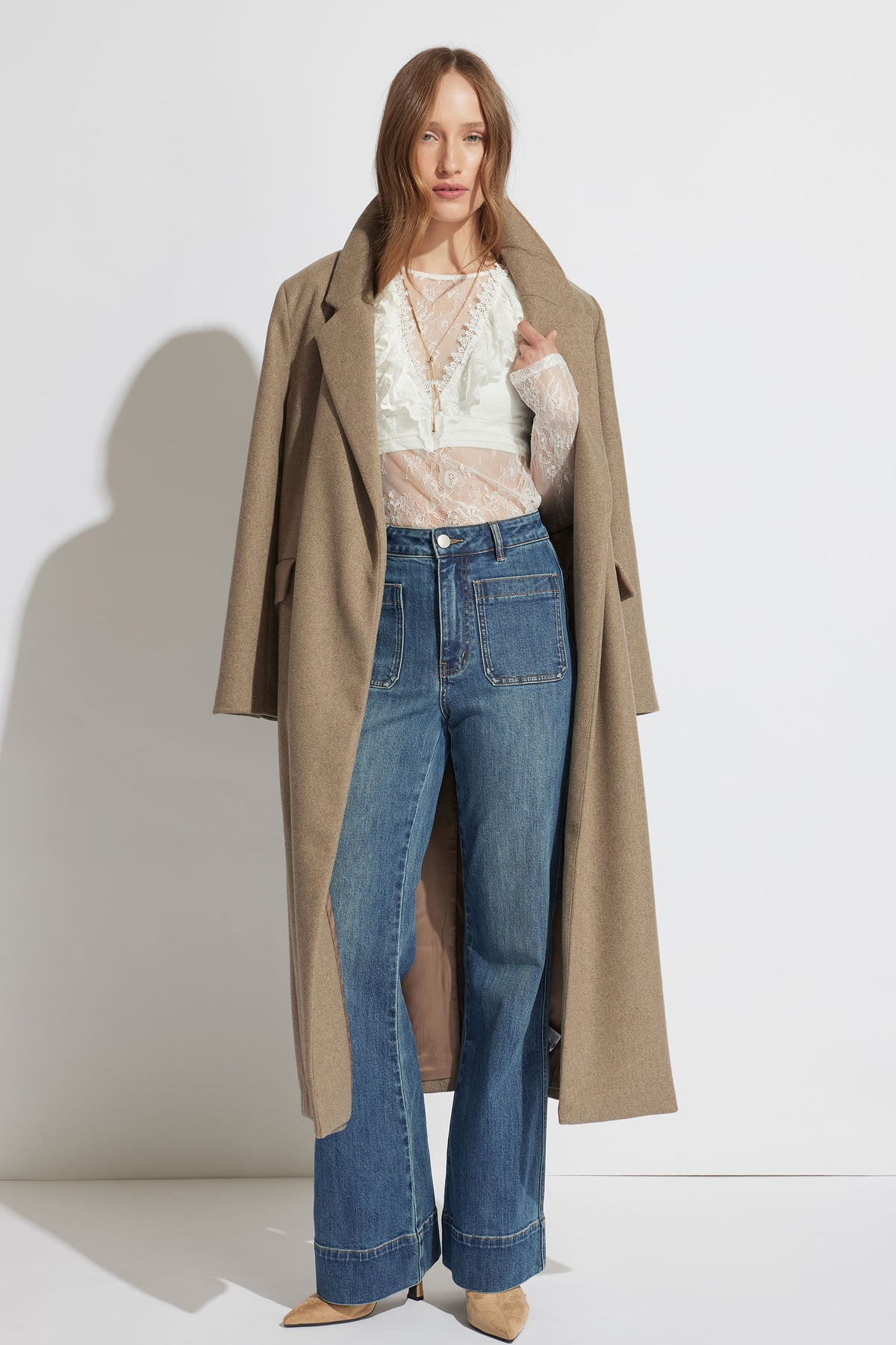 Woman wearing a sheer white lace top layered with a beige long coat and high-waisted wide-leg blue jeans