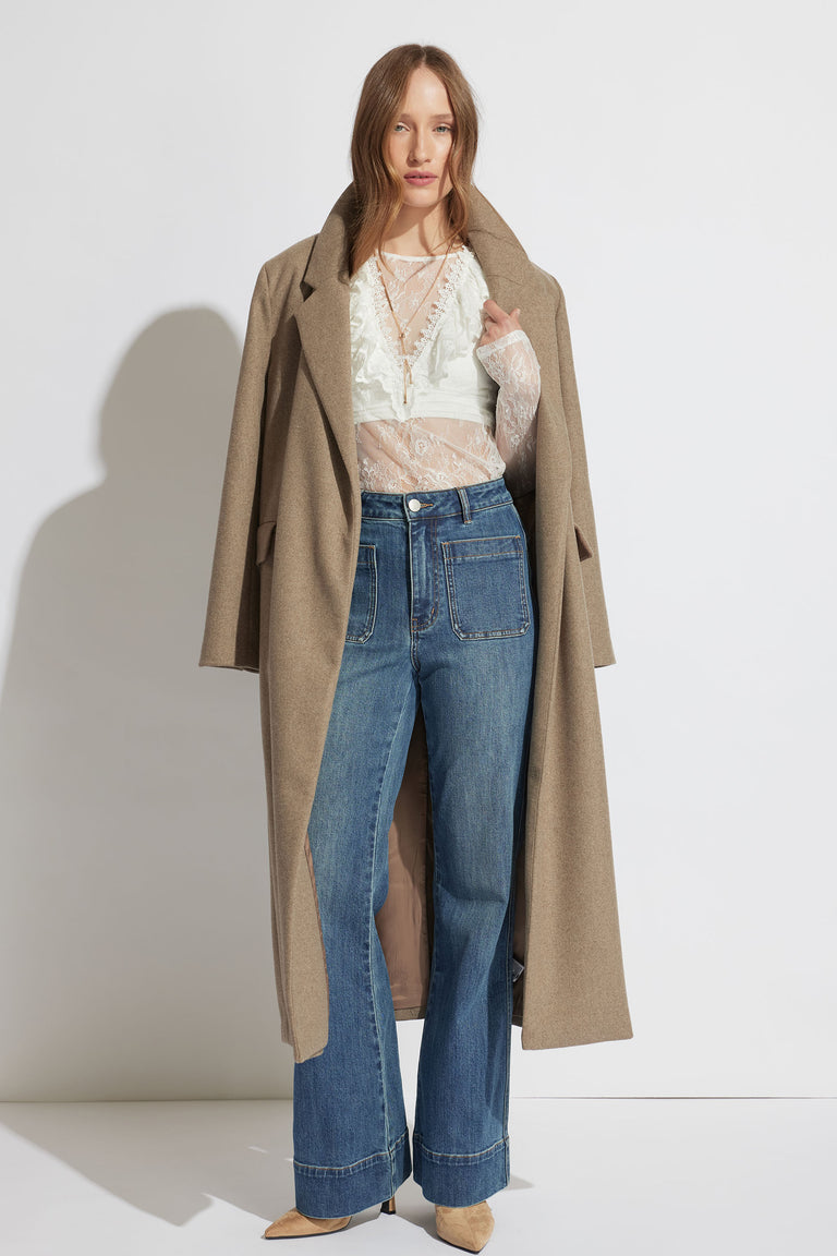 Woman wearing a sheer white lace top layered with a beige long coat and high-waisted wide-leg blue jeans