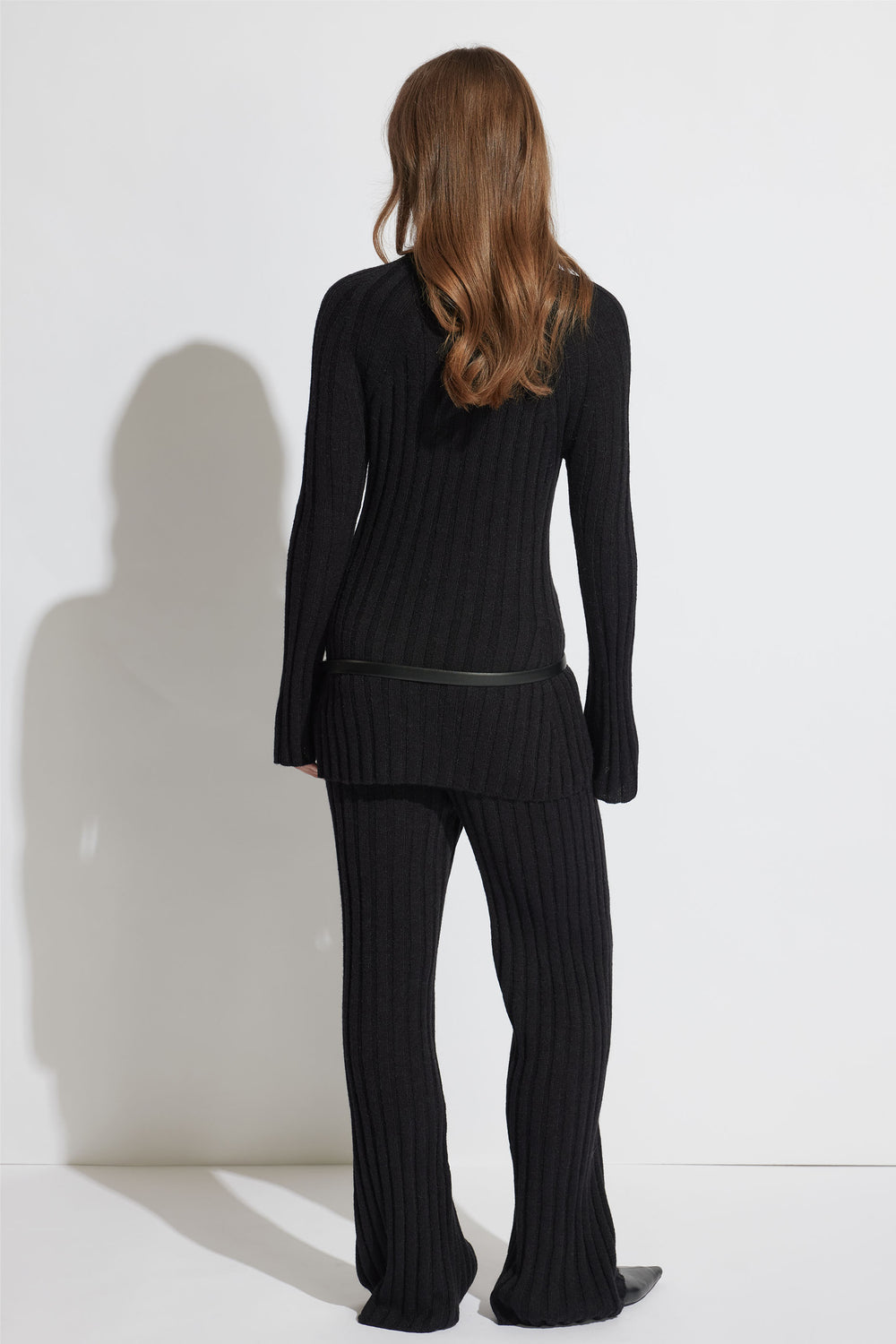 Woman with loose wavy hair wearing black rib-knit pants and a matching long sweater with a slim belt, back view.