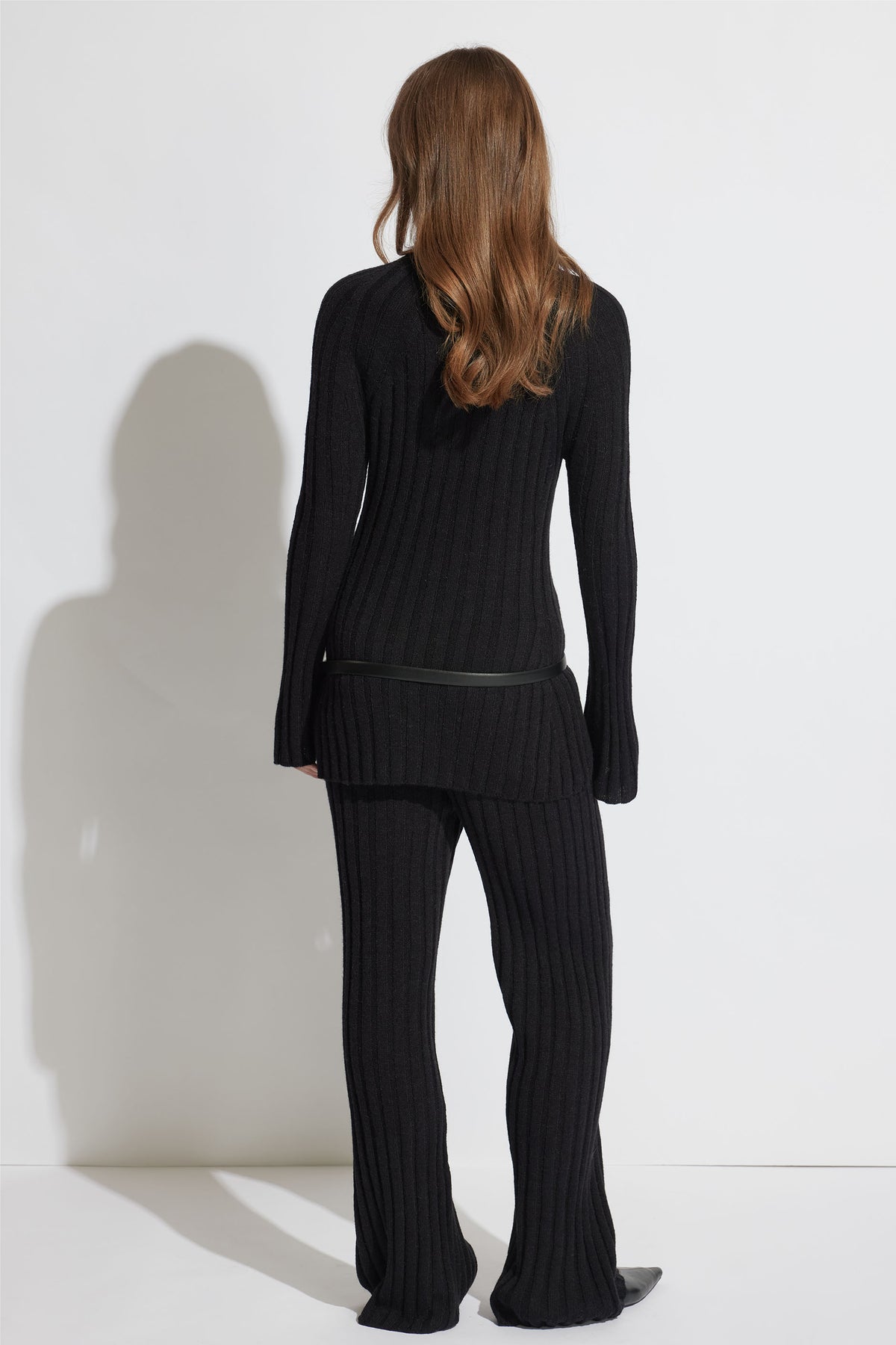 Woman with loose wavy hair wearing black rib-knit pants and a matching long sweater with a slim belt, back view.