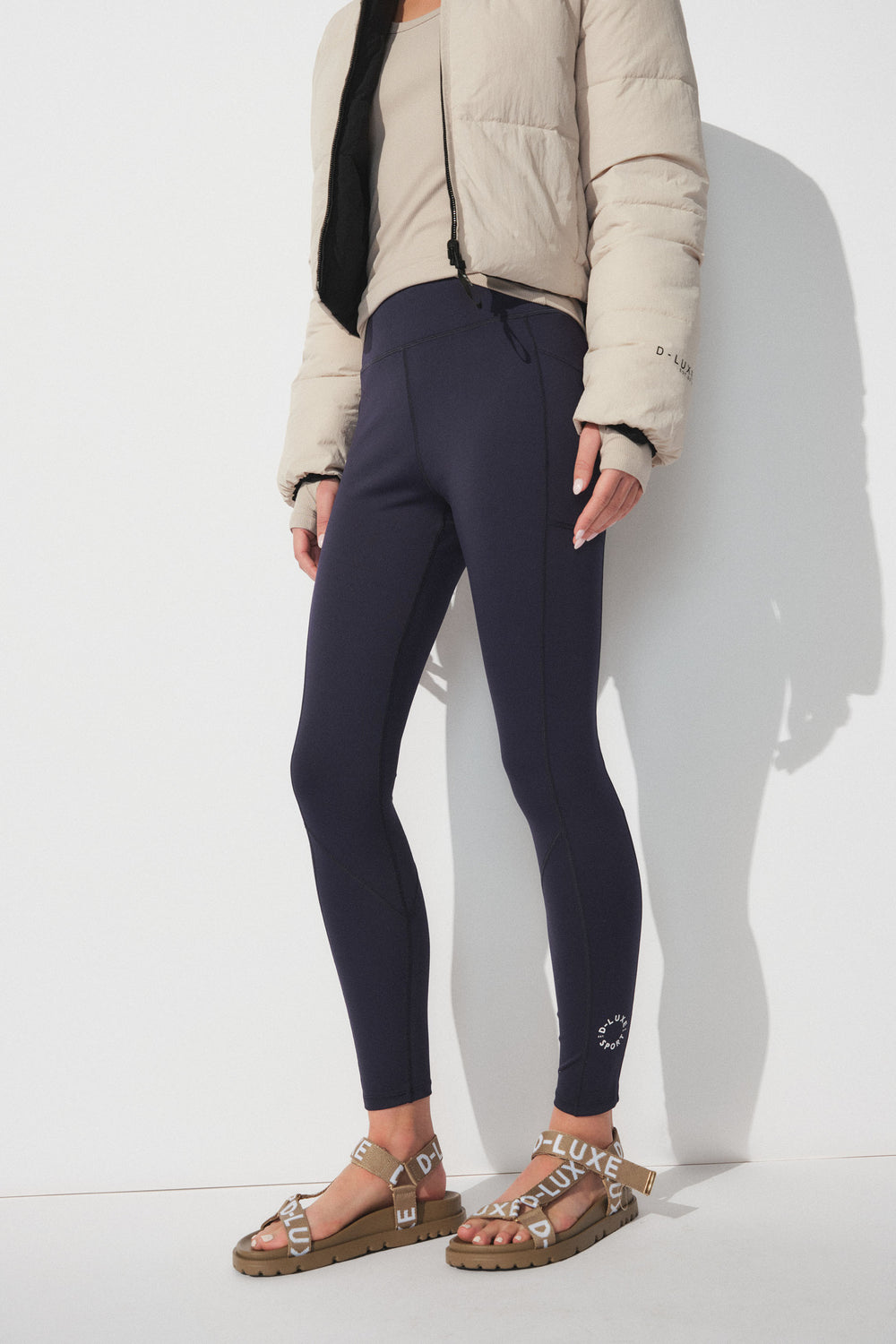 Woman wearing navy D-Luxe leggings styled with a beige crop puffer jacket and branded strap sandals