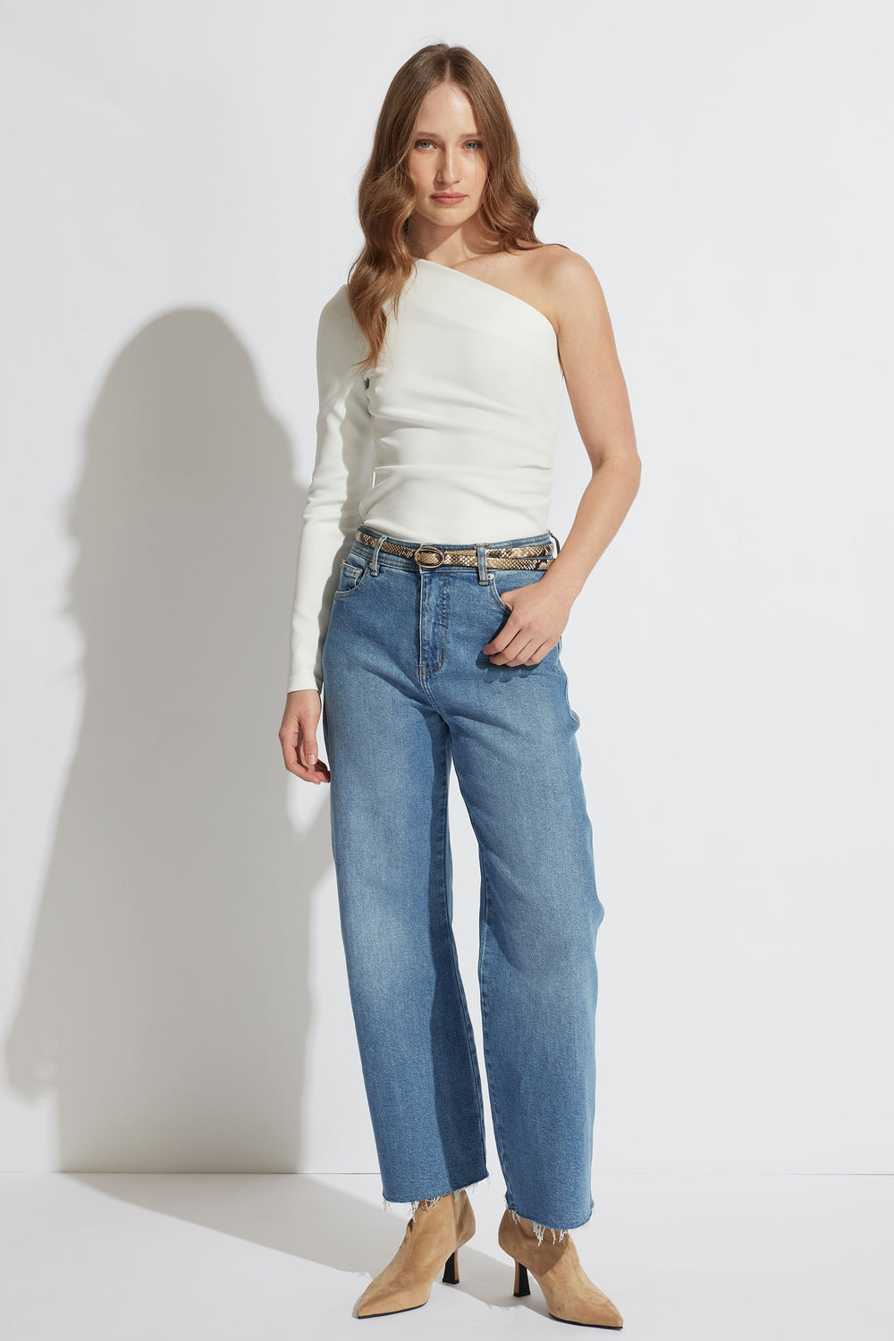 Woman wearing an ivory one-shoulder top paired with high-waisted straight-leg blue jeans and tan ankle boots.