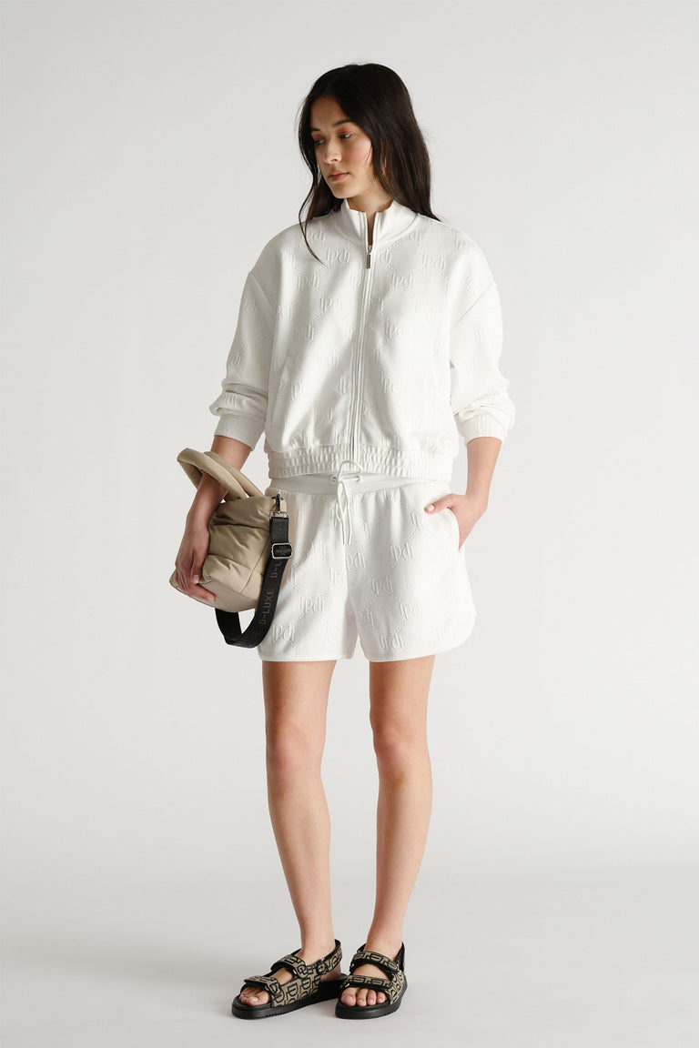 Woman wearing an embossed white sweat short paired with a matching zip-up jacket and black patterned sandals, holding a beige bag.