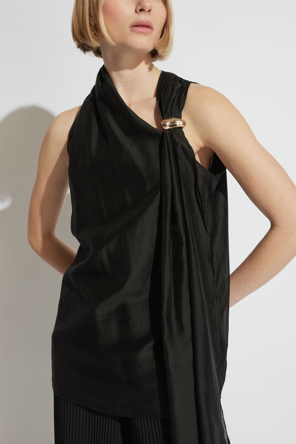 Woman wearing a black draped top with a gold-tone ring detail over one shoulder, styled with black pants.