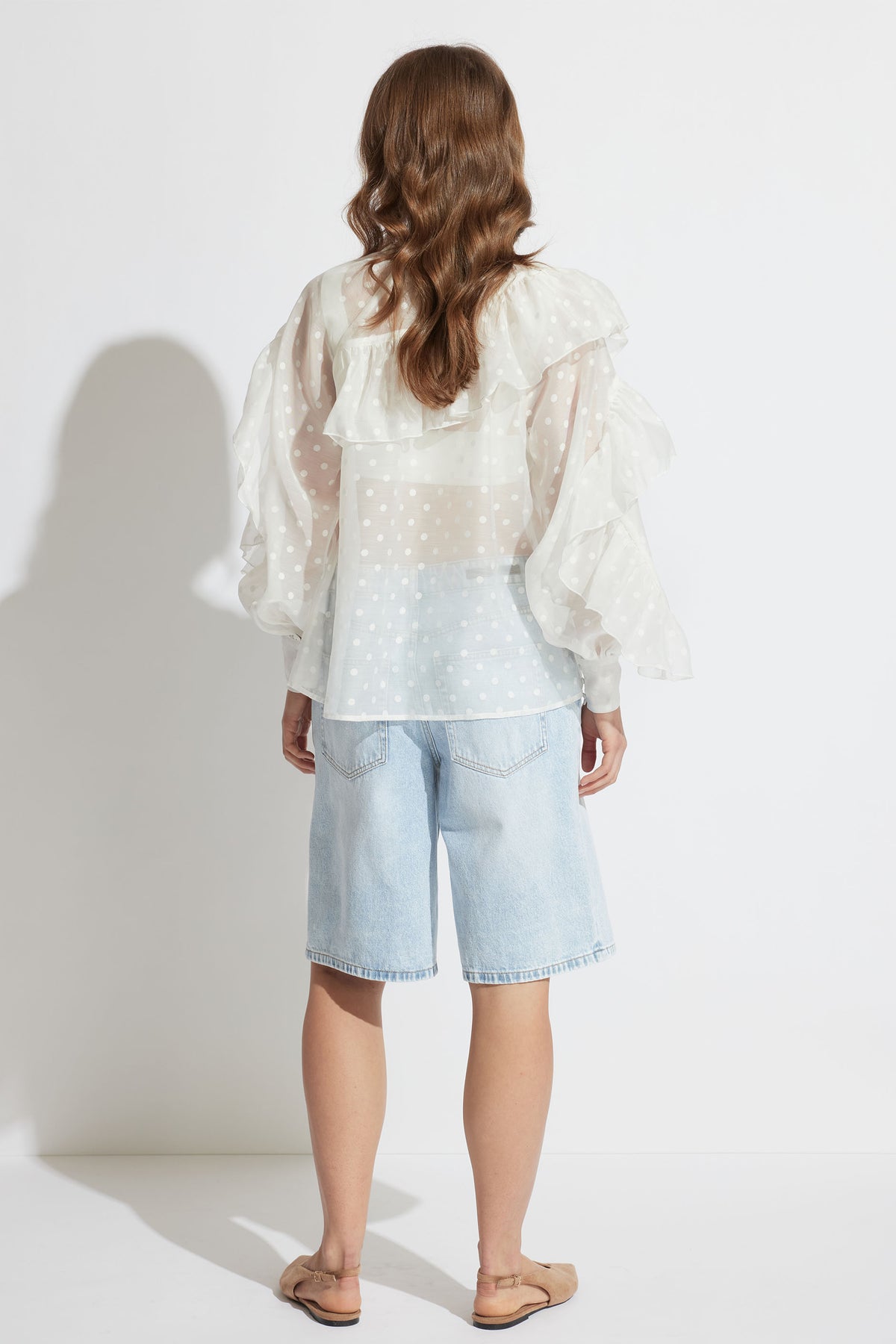 Back view of woman wearing a sheer white spot ruffle top with voluminous sleeves and light denim long shorts