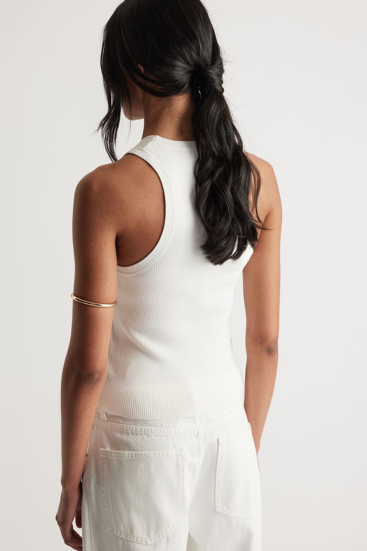 Back view of a woman wearing an ivory ribbed knit racerback tank paired with white high-waisted pants and a gold arm cuff.