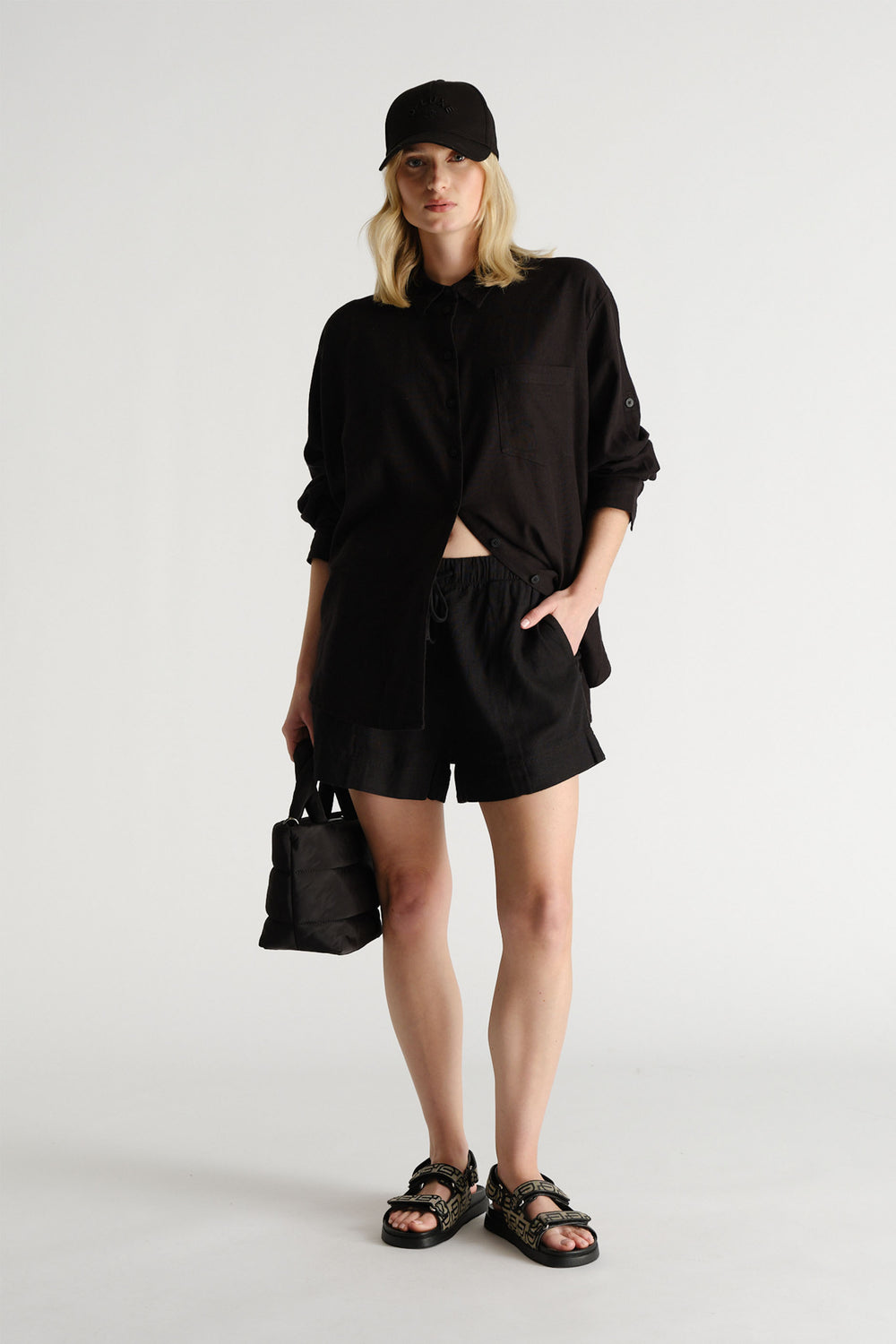 Portrait of a model wearing black D-Luxe Linen Blend Short with matching top, cap, and sandals in gallery position 1.