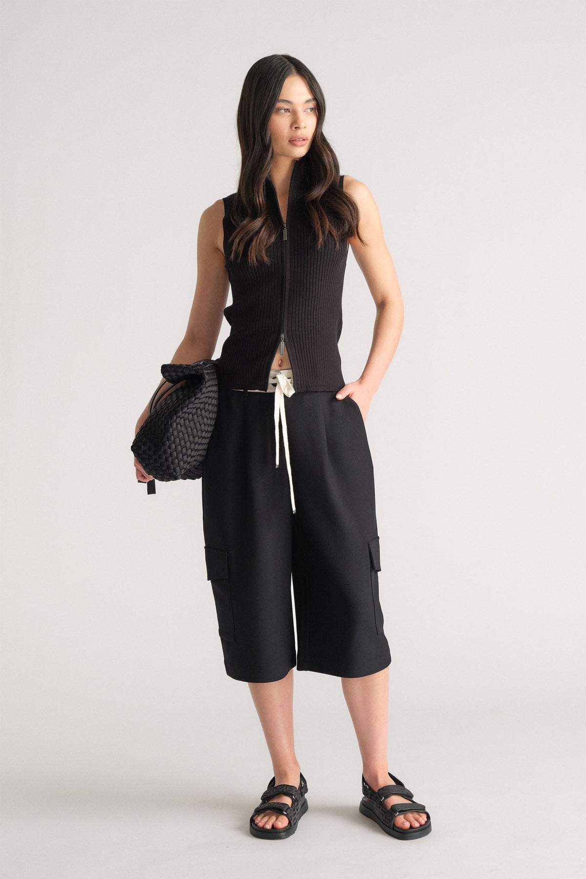 Model wearing black D-Luxe Logo Waistband Short with cargo pockets in portrait gallery position 1, styled with black sleeveless top and sandals