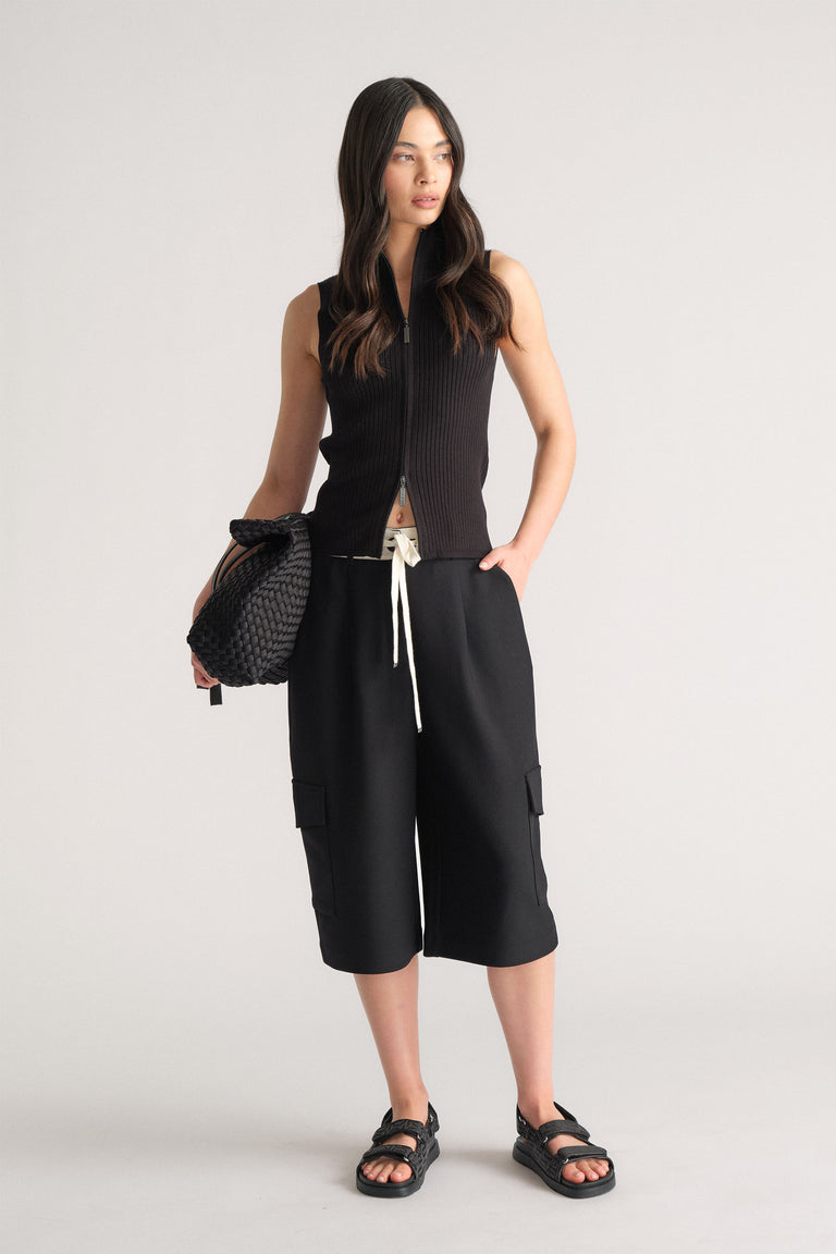 Model wearing black D-Luxe Logo Waistband Short with cargo pockets in portrait gallery position 1, styled with black sleeveless top and sandals