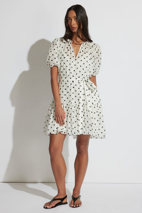 Woman wearing a white mini dress with black polka dots, puffed short sleeves, and a tied waist detail paired with black sandals.