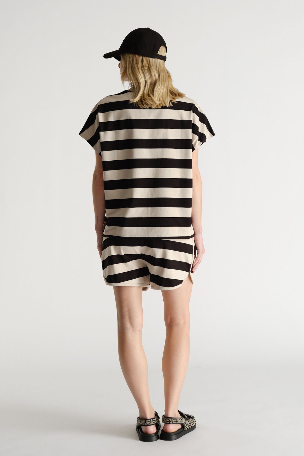Woman wearing a black and shell color bold striped short set with a black cap and patterned sandals, shown from behind.