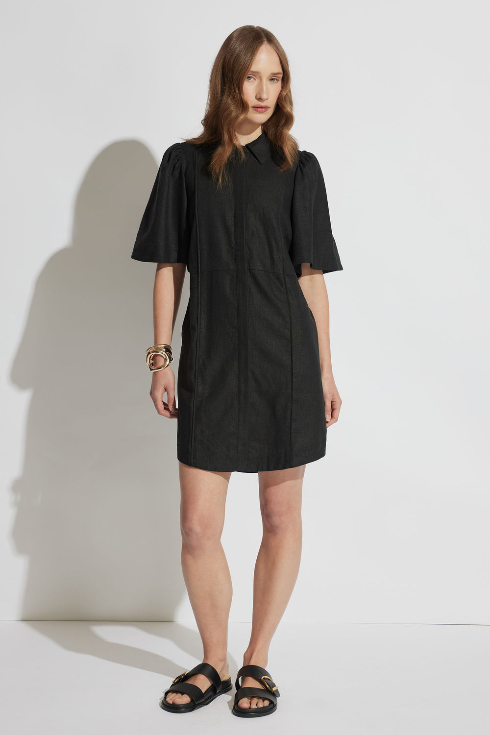 Woman wearing a black mini shirt dress with puffed sleeves, accessorized with bangles and black sandals, standing against white wall.