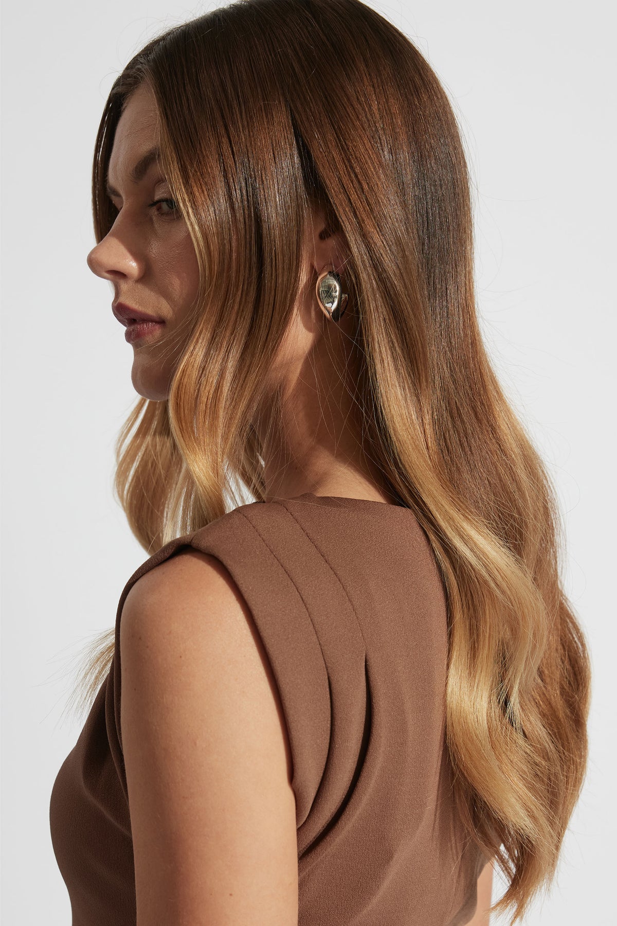 Profile of woman with long wavy hair wearing silver abstract hoop earrings and a sleeveless brown top against a plain background