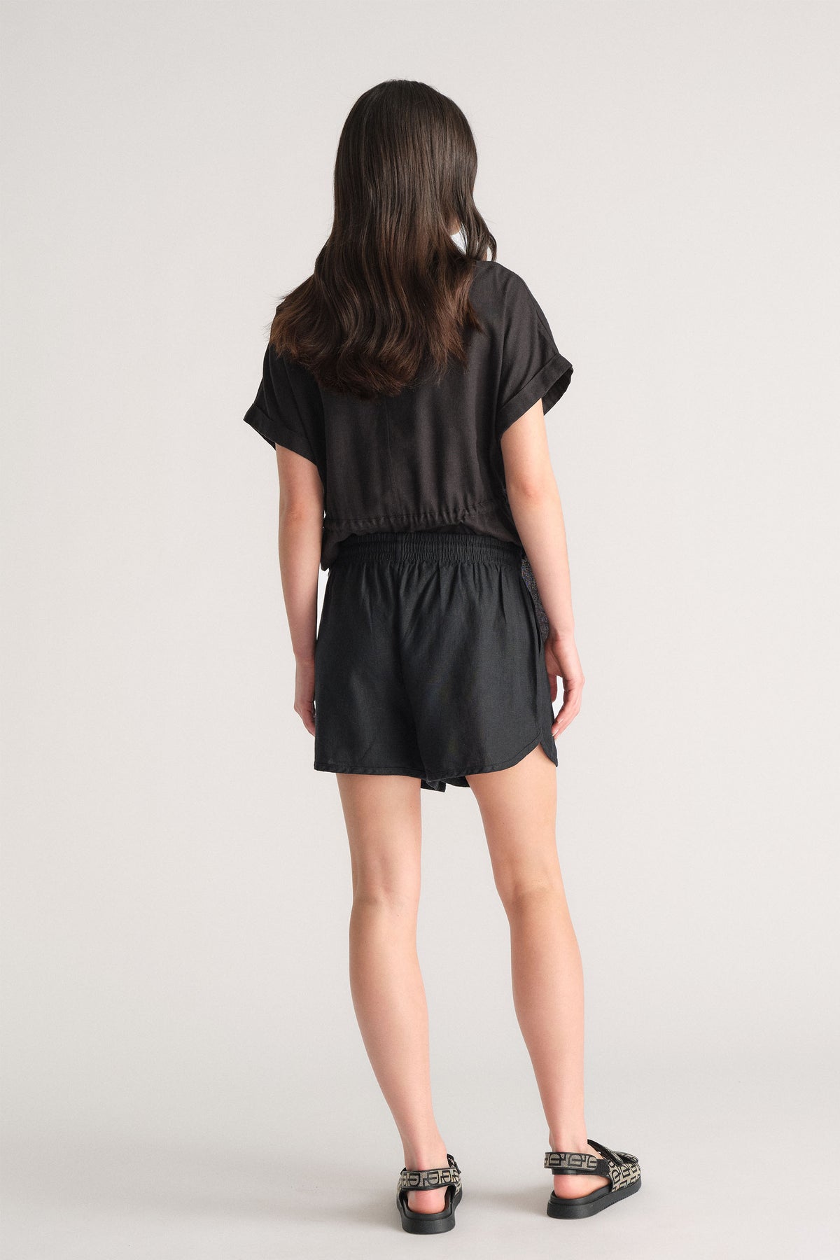 Girl wearing black linen blend shorts with an elastic waistband paired with a loose black short-sleeve top, shown from behind.