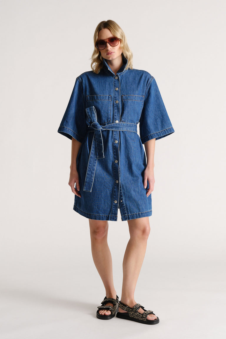 Woman wearing a belted medium blue denim dress with wide sleeves and front buttons paired with patterned sandals and sunglasses.