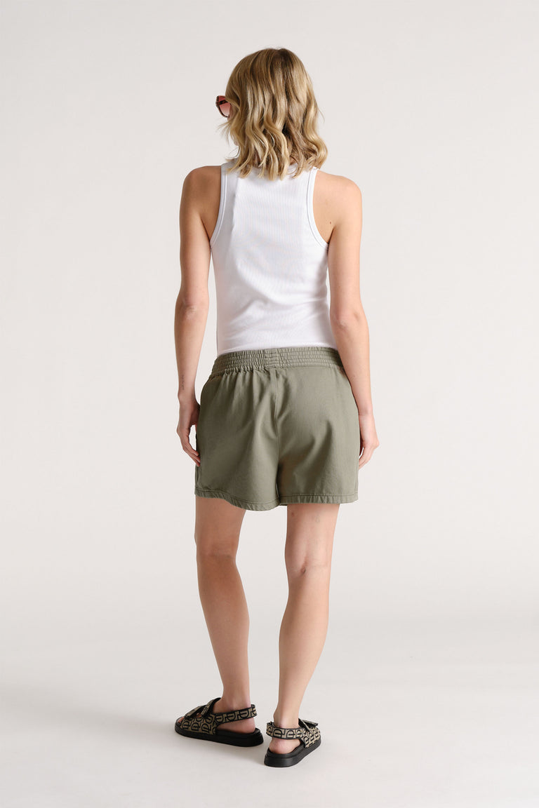 Woman wearing olive green pull-on shorts with an elastic waistband, paired with a white ribbed tank top and patterned sandals.
