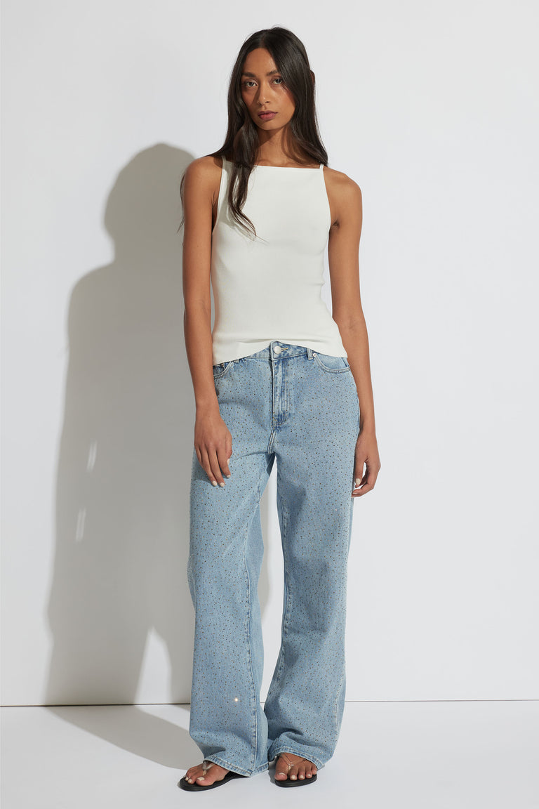 Woman wearing an ivory square neck tank paired with loose light blue denim jeans and black sandals in a minimalist setting.