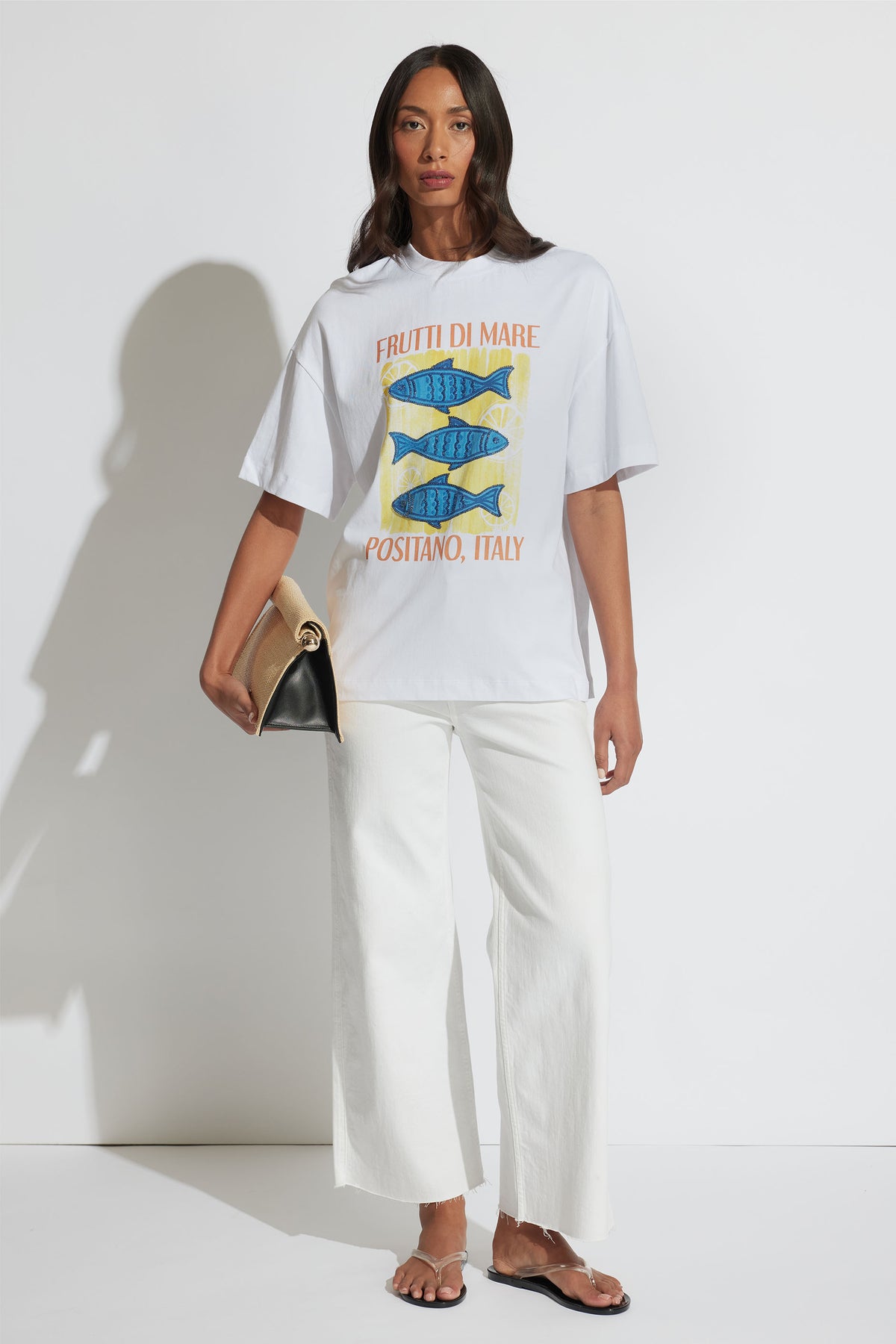 Portrait of model in Coastal Beaded Tee with blue fish graphic, paired with white pants and tan clutch, gallery position 2