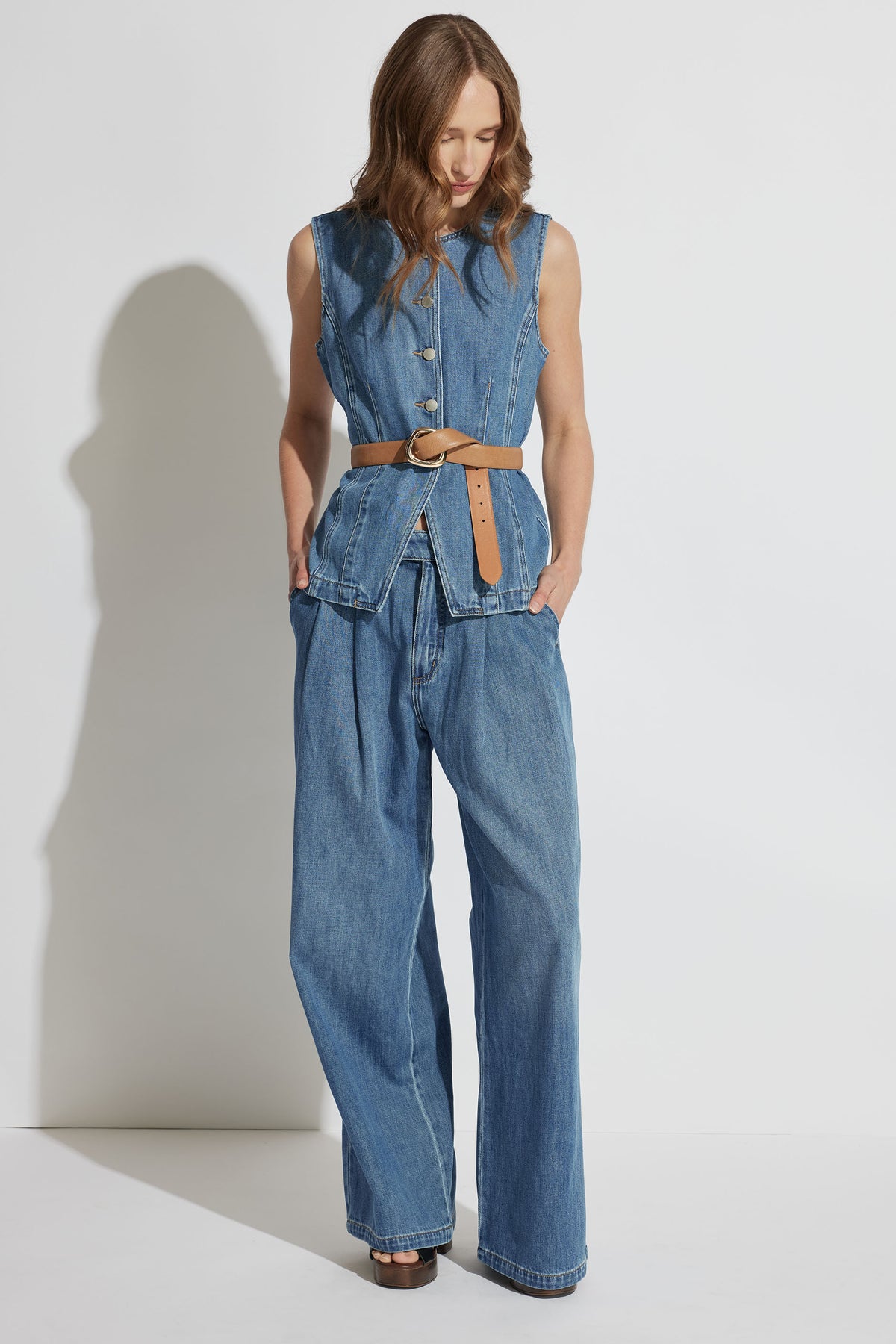 Woman wearing a sleeveless denim vest with large buttons and pleated wide-leg denim jeans styled with a tan belt