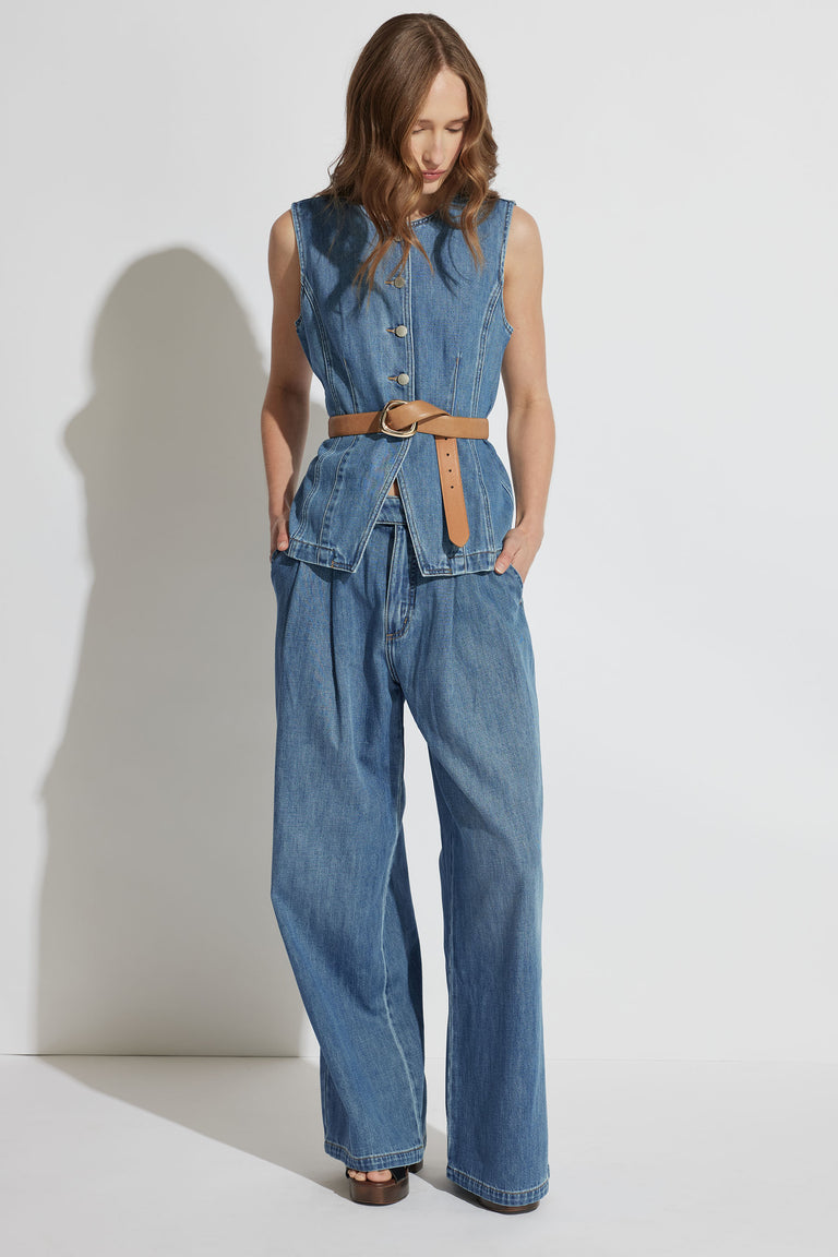 Woman wearing a sleeveless denim vest with large buttons and pleated wide-leg denim jeans styled with a tan belt