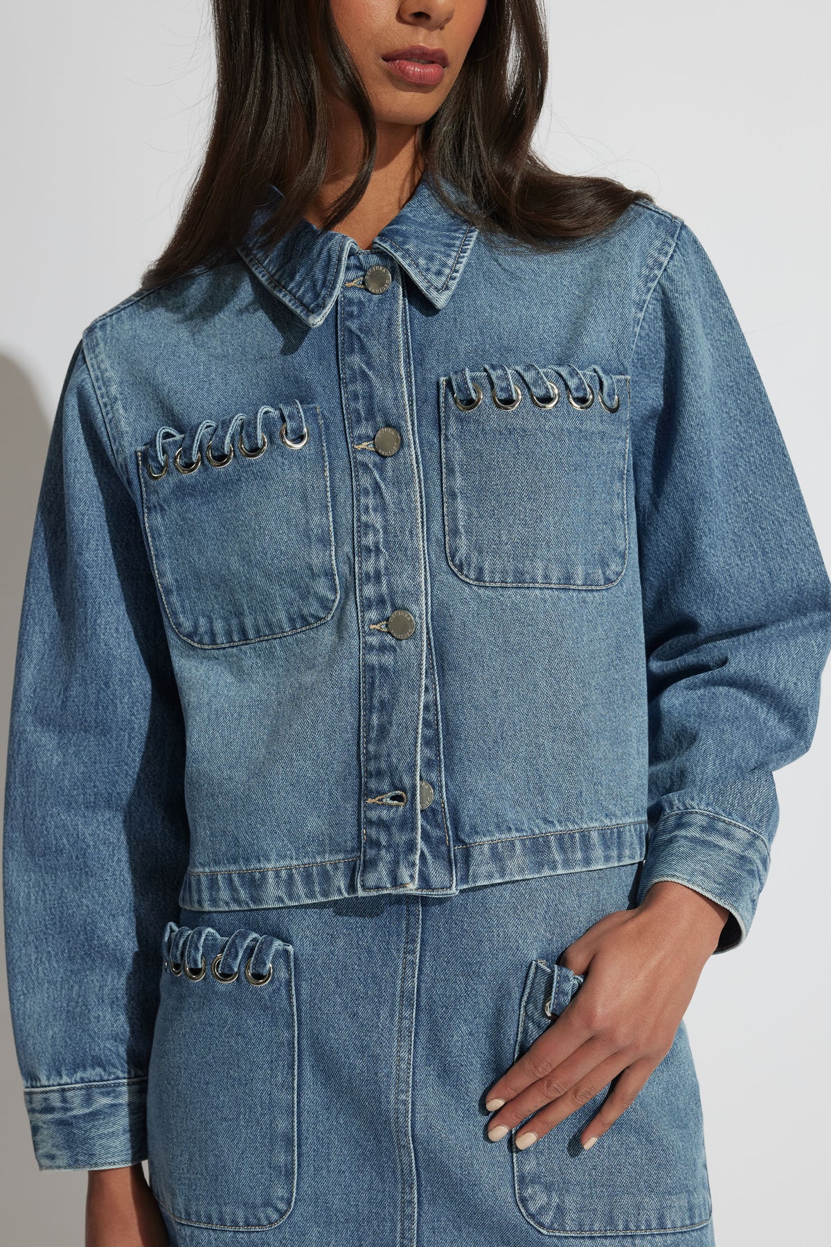 Woman wearing a medium blue denim jacket with metal ring detailing on front pockets and matching skirt, casual style.