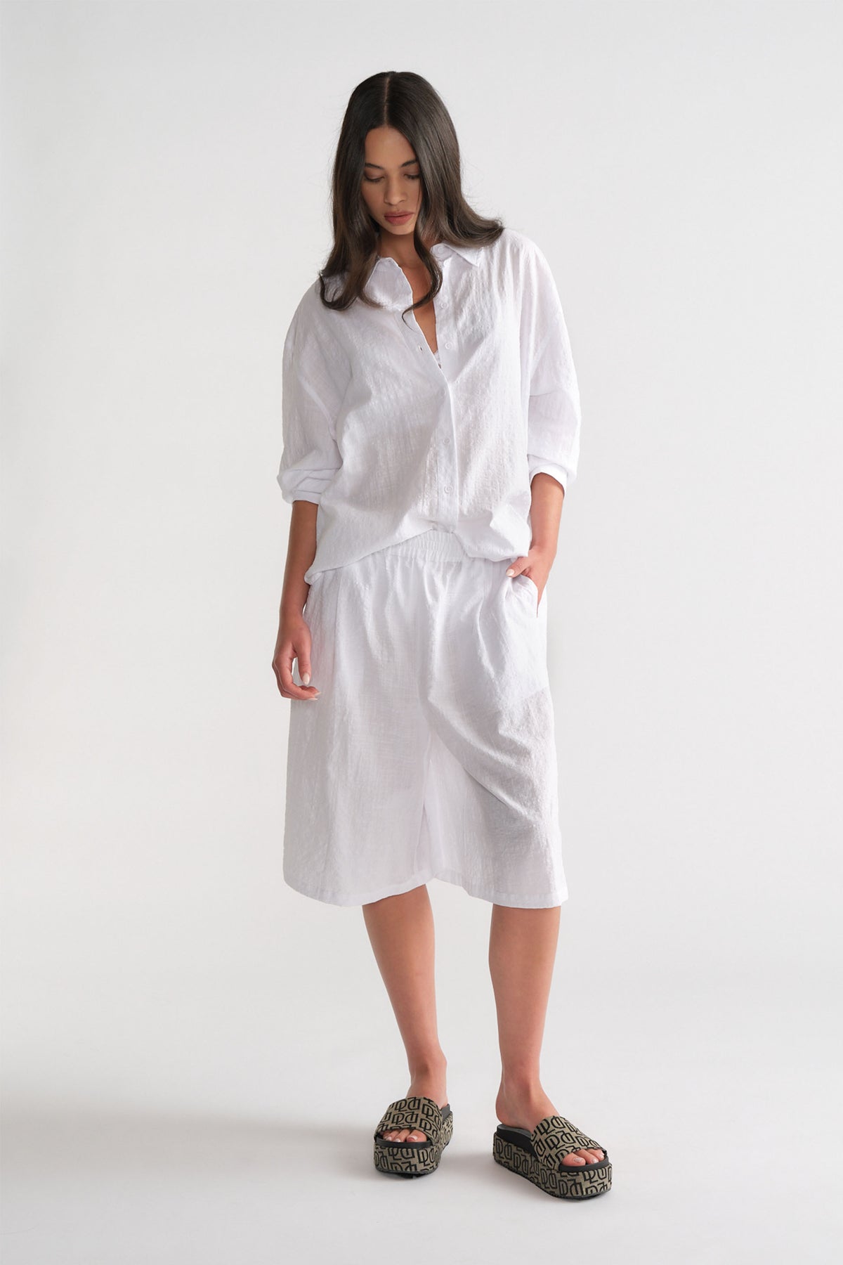 Woman dressed in a white relaxed linen button-up shirt and matching Bermuda shorts, styled with patterned platform sandals.