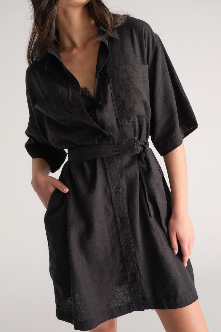 Woman wearing a black linen blend short dress with a belted waist and button-up front, styled with lace detail underneath.