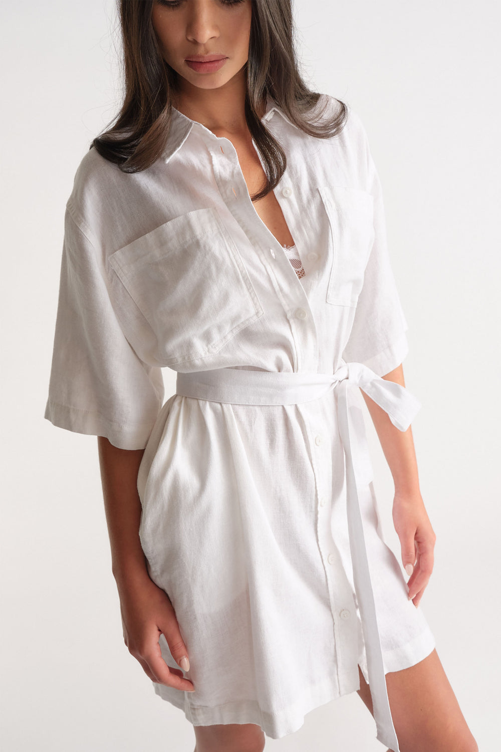 Woman wearing a white linen blend short dress with large front pockets and a tied waist belt for a relaxed fit