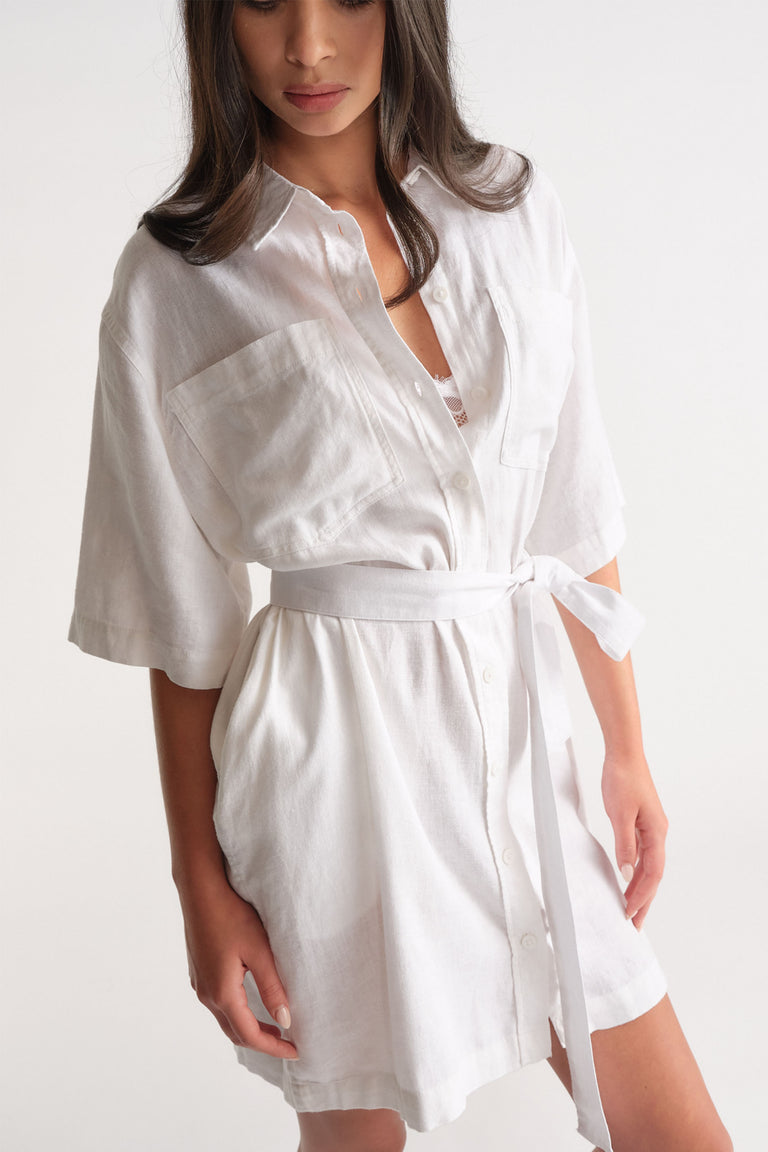 Woman wearing a white linen blend short dress with large front pockets and a tied waist belt for a relaxed fit