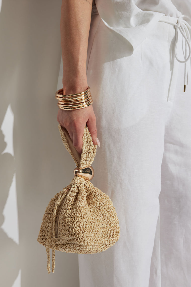 Hand wearing stacked gold bangles holding a small woven straw bag, styled with relaxed white linen pants.