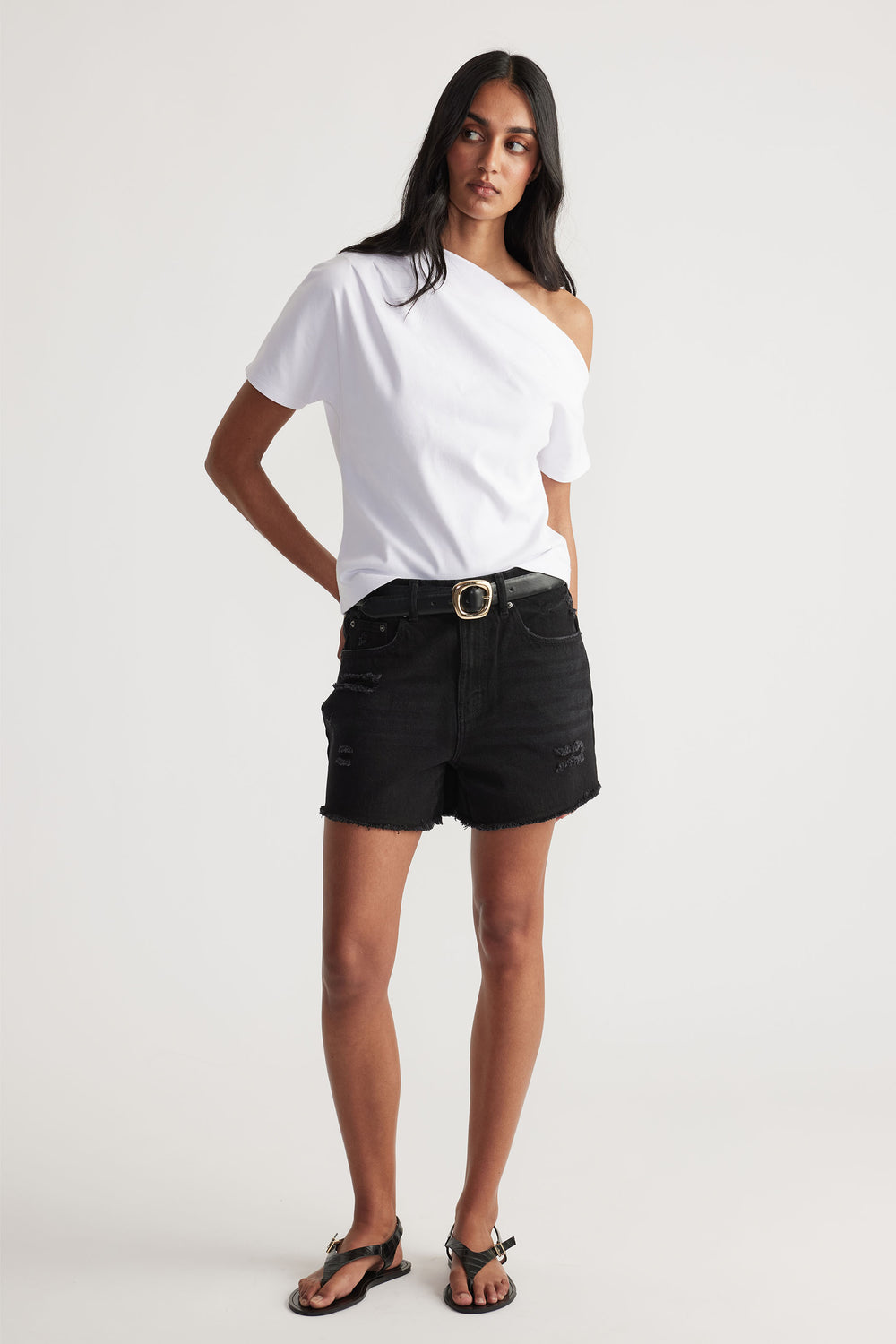 Woman wearing black distressed denim shorts with belt, paired with white one-shoulder casual top and black sandals.