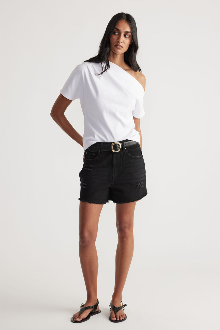 Woman wearing black distressed denim shorts with belt, paired with white one-shoulder casual top and black sandals.