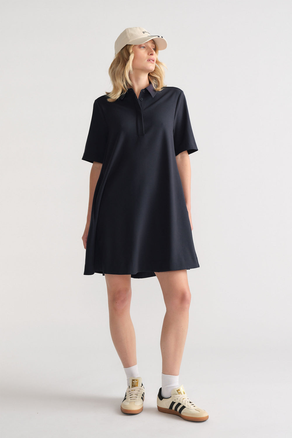 Woman wearing a relaxed black polo dress with short sleeves styled with a beige baseball cap and casual sneakers