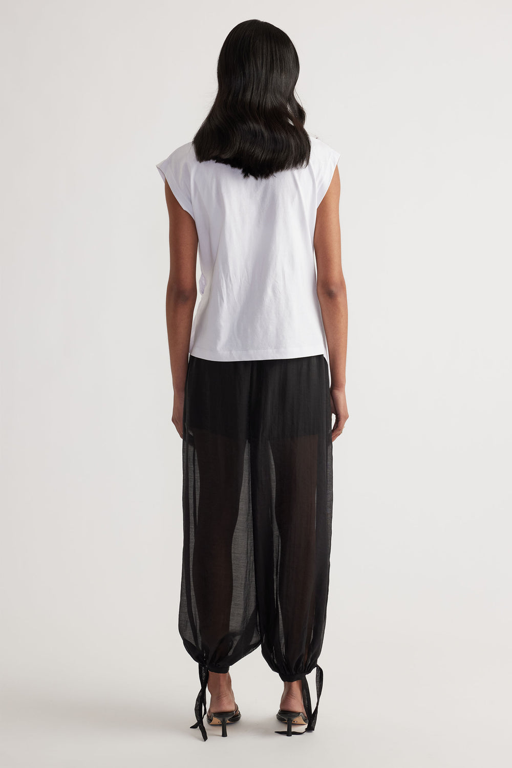 Woman seen from behind wearing a white sleeveless top paired with sheer black wide-leg pants featuring ankle ties and heels