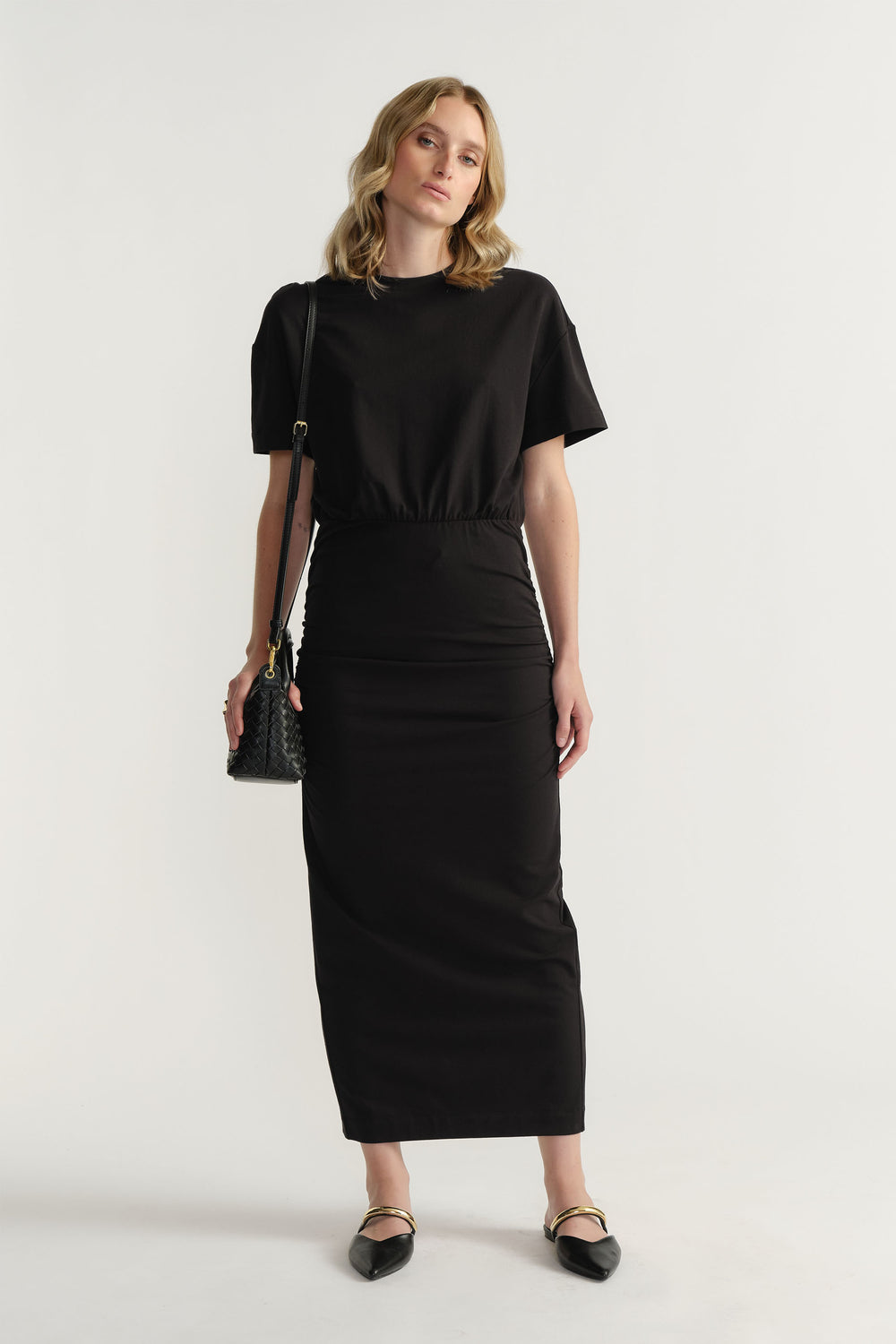 Woman wearing a sleek black ruched midi dress with short sleeves, paired with a small textured shoulder bag and pointed flats
