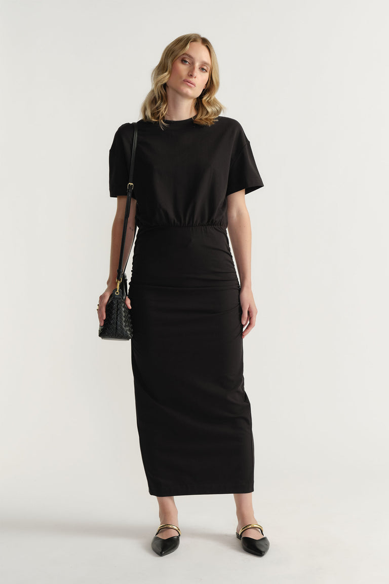 Woman wearing a sleek black ruched midi dress with short sleeves, paired with a small textured shoulder bag and pointed flats