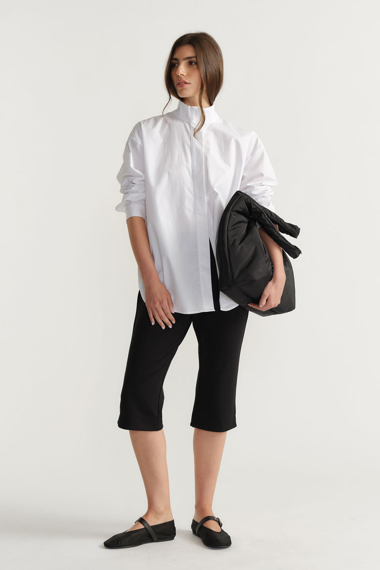 Woman wearing a relaxed white button-up shirt and black cropped pants, holding a large black puffer bag in one arm.