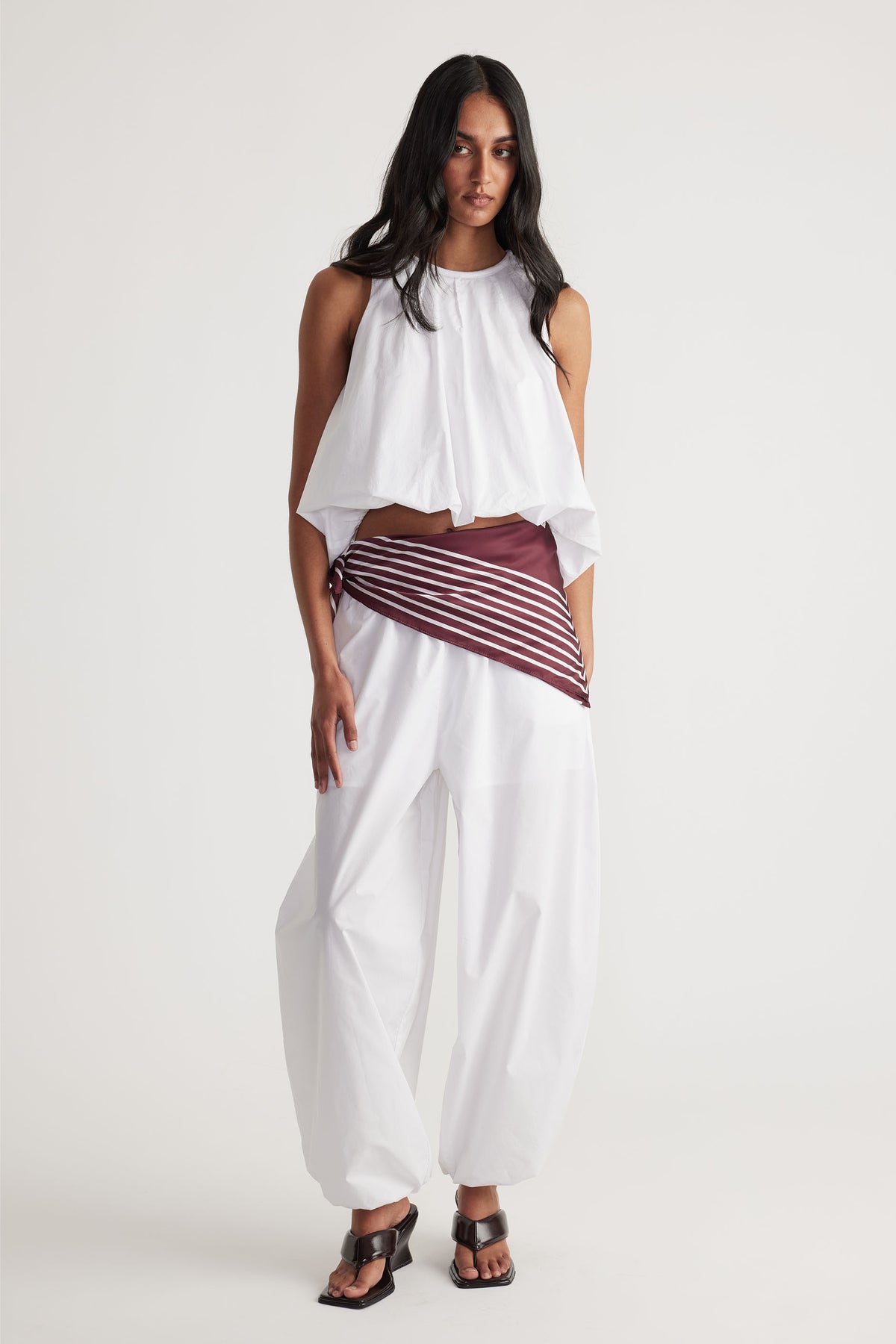 Woman wearing loose white cotton balloon pants paired with a sleeveless white draped top and brown striped belt detail