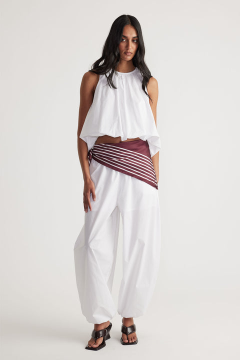 Woman wearing loose white cotton balloon pants paired with a sleeveless white draped top and brown striped belt detail