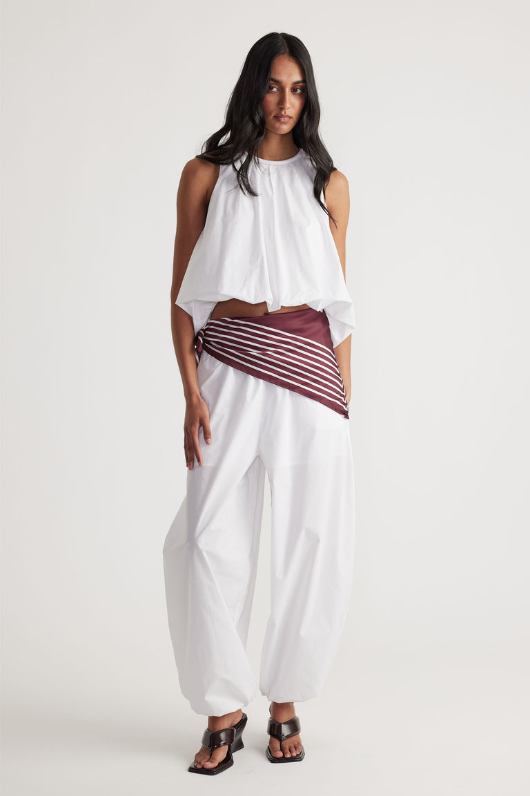 Woman wearing loose white cotton balloon pants paired with a sleeveless white draped top and brown striped belt detail